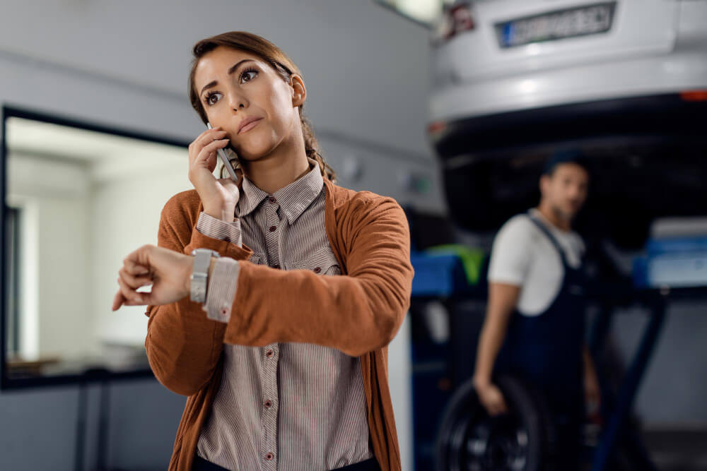 Eine Frau telefoniert und schaut ungeduldig auf die Uhr, während im Hintergrund ein Mechaniker einen Reifen in der Hand hält