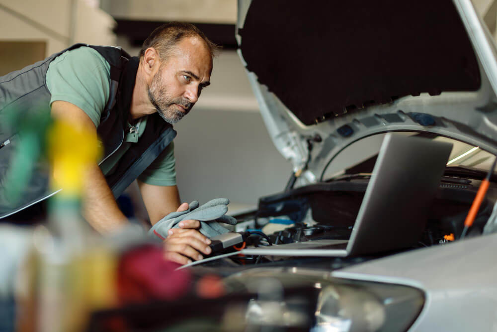 Ein Mechaniker erstellt am Laptop ein Schadensgutachten, während er in der Werkstatt unter der Motorhaube eines Autos arbeitet